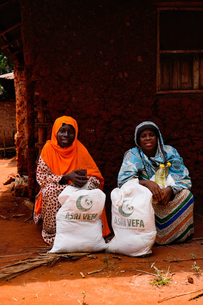 services-04 Two women sit by a mud house with relief bags, showing community aid.